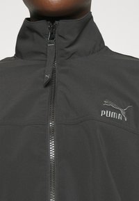Svart zip-up jacka med ståkrage, tillverkad av slätt tyg, med en subtil Puma-logotyp i grått på vänster bröst och horisontell sömnadsdetalj.
