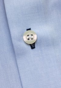 Close-up van een witte knoop genaaid met witte draad op lichtblauwe stof, bevestigd door een rechthoekig donkerblauw knoopsgat.