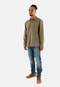 Camisa de botón verde oliva con mangas largas, combinada con jeans de color azul claro y zapatillas altas marrones. Diseño simple, sin patrones notables.
