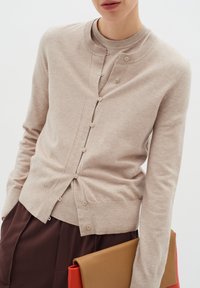 Beiger Cardigan mit Rundhalsausschnitt, Vorderknöpfen und feiner Stricktextur. Kombiniert mit braunen Hosen und einer farbigen Klapp-Clutch.