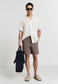 Kurzärmliges, gestreiftes cremefarbenes Hemd mit braunen Akzenten, kombiniert mit hellbraunen Shorts. Beigefarbene Slippers und eine dunkelblaue Tragetasche.