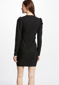 Robe mini noire à côtes avec manches longues, présentant un motif texturé et un subtil éclat, avec un resserrement à la taille et des accentuations de manches bouffantes.