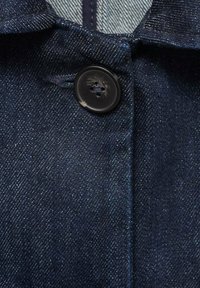 Temno modra denim tkanina z diagonalnim tkanjem, opremljena z črnim plastičnim gumbom s štirimi luknjami. Material ima gladko teksturo in subtilen lesk.