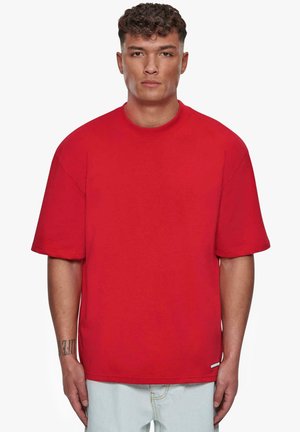 T-shirt surdimensionné en coton rouge avec des manches courtes et un col rond. Présente une étiquette décorative près de l'ourlet. Couleur unie, sans motifs.