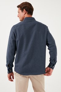 Camicia button-up a maniche lunghe di colore blu scuro con un tessuto texturizzato, colletto a punta e polsini con bottoni. La camicia è indossata sopra pantaloni di colore chiaro.
