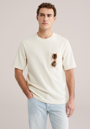 Crèmekleurig shirt met korte mouwen van getextureerde stof en een borstzak, met een bruine zonnebril erin. Gecombineerd met lichtblauwe spijkerbroek.