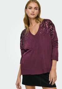 V-Ausschnitt, oversized Strickpullover in tiefem Pflaumenton mit schwarzen und pinken Paillettenverzierungen an den Schultern. Getragen über einem schwarzen Cordminirock.