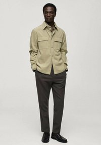 Olijfgroene corduroy shirt met twee borstzakken, knoopsluiting en lange mouwen, gecombineerd met donkerbruine broek en zwarte schoenen.
