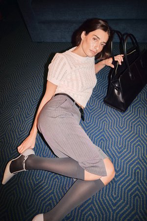 Svart tote-väska, ljus stickad topp, grå randiga byxor, grå knähöga strumpor och beige spetsiga skor. Texturerad blå matta med zigzag-mönster.