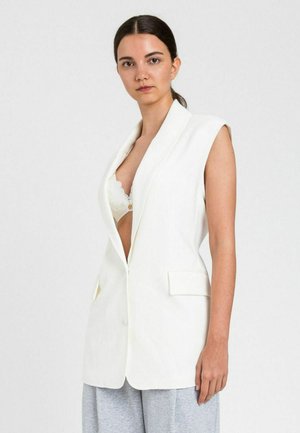 Giovane donna con capelli scuri in blazer bianco senza maniche sopra un reggiseno bianco in pizzo e pantaloni grigio chiaro, espressione neutra, sfondo semplice.