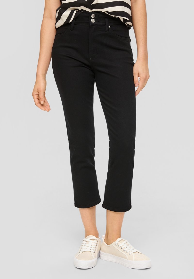 Femme portant un pantalon flare court noir, des baskets beiges et un haut rayé noir et blanc sur un fond uni.
