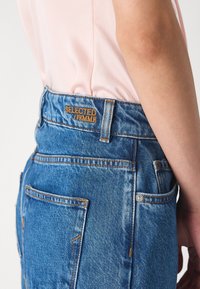 Zili denimi džinsi ar augsto jostasvietu, ar zeltā izšūtu "SELECTED FEMME" etiķeti un kontrastējošām oranžām šuvēm.