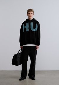 Μαύρο φούτερ με κουκούλα με μεγάλο ακουα χρώματος λογότυπο "HU", συνδυασμένο με φαρδιά μαύρα παντελόνια και μαύρα παπούτσια. Κρατάει μια μαύρη τσάντα. Απλός, μοντέρνος σχεδιασμός.