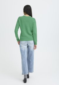 Mujer con cabello largo y oscuro, vistiendo un suéter verde texturizado, jeans de pierna ancha azul claro, calcetines grises y zapatos negros, vista desde atrás.