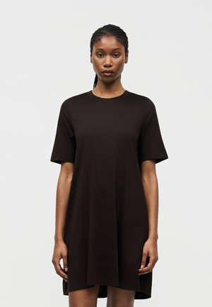 Γυναίκα με πλεξούδες, φορώντας ένα φαρδύ μαύρο φόρεμα με κοντά μανίκια, όρθια μπροστά σε απλό λευκό φόντο.