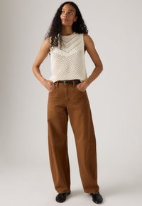 Vrouw met lang krullend haar, gekleed in een witte mouwloze haaktop, bruine wijde broek, zwarte riem en zwarte schoenen, staande met haar handen in de zakken.