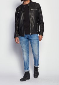 Giacca di pelle nera con colletto alto, dotata di zip frontali e due tasche sul petto. Indossata sopra una camicia nera e jeans blu.