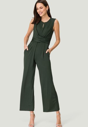 Une combinaison vert foncé avec un corsage ajusté, des jambes larges et des poches. Elle présente un détail en trou de serrure au niveau du col et un design de taille drapée.