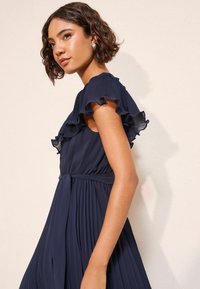 Tumesinine volditud kleit volangvarrukatega ja vöökohaga. Sile kangas elegantse langevusega, mis rõhutab kontuure ja liikumist.