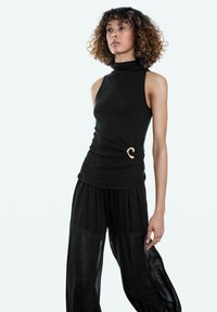 Svart ärmlös polotröja med figurnära passform och guldfärgat spänndetalj vid midjan, kombinerad med transparenta svarta byxor med vida ben.