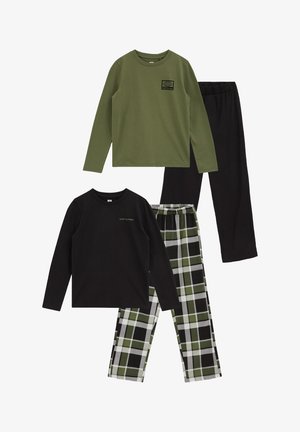 Garçon aux cheveux bouclés portant une chemise verte à manches longues avec un patch noir, un pantalon noir et des chaussettes blanches sur fond uni.