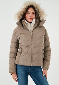 Giubbotto imbottito marrone con cappuccio foderato in pelliccia, chiusura con zip e due tasche laterali, abbinato a un maglione a coste di colore chiaro e jeans blu.