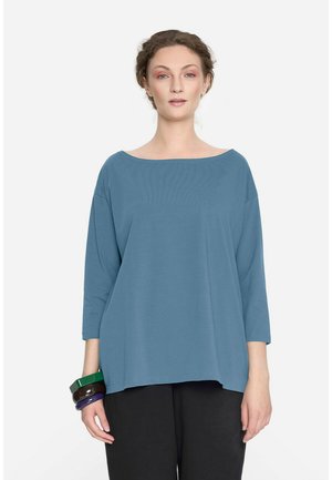 Kobieta z plecionymi włosami, ubrana w luźną niebieską bluzkę z rękawami do łokci, czarne spodnie i kolorowe bransoletki na lewej ręce.