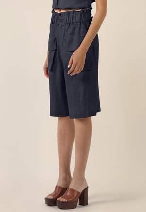 Mujer con pantalones cortos azul marino hasta la rodilla con grandes bolsillos delanteros y un top negro suelto y corto, combinado con sandalias marrones de tacón alto.