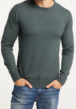Homme portant un pull col rond vert foncé et un jean bleu, les mains posées décontractées dans les poches, sur fond blanc.