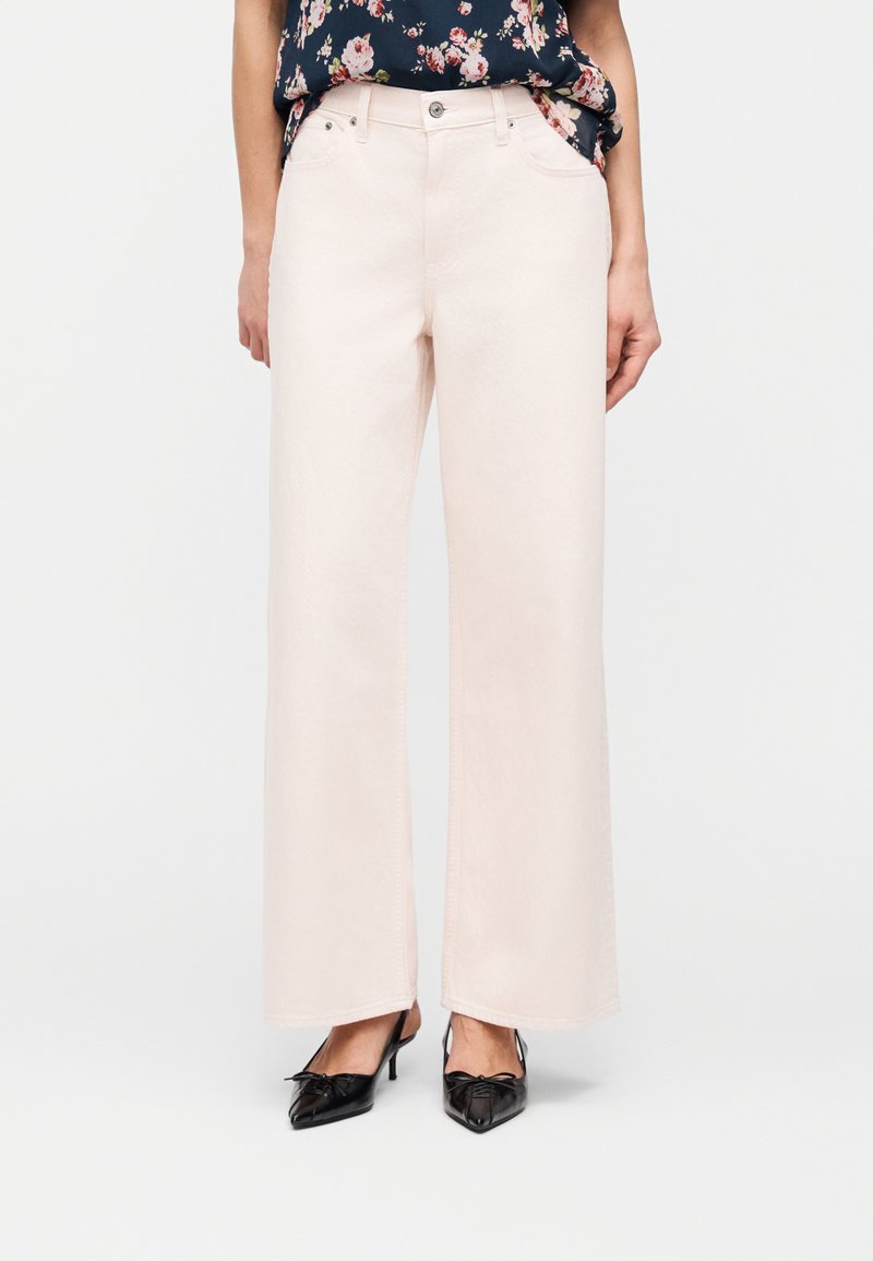 Γυναίκα που φοράει ροζ παντελόνι ψηλής μέσης και φαρδιάς γραμμής, με μαύρα μυτερά παπούτσια τύπου kitten heels και φλοράλ αμάνικη μπλούζα.