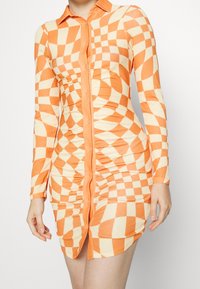Abito a maniche lunghe a quadri arancioni e panna con chiusura frontale a bottoni, caratterizzato da una texture arricciata lungo i lati e una silhouette aderente.