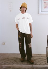 Biała oversize koszulka z logo Toyota, zestawiona z ciemnymi, luźnymi spodniami z naszywkami i haftami, noszona z żółtą dzianinową czapką.