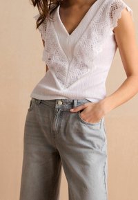 Haut blanc avec bordure en dentelle à motifs, décolleté en V et tissu texturé, associé à un jean taille haute gris clair avec poches avant.