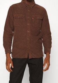 Braunes Cordhemd mit durchgehender Knopfleiste, zwei Brusttaschen und einem Hemdkragen. Ausgezeichnet durch eine strukturierte Stoffqualität und ein dezentes Markenetikett.