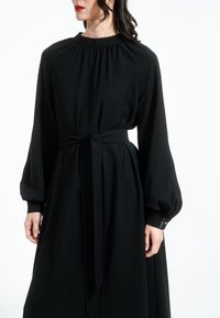 Crna haljina dugih rukava od mekog materijala, s nabranim izrezom, pojasom u struku i rukavima s manžetama i detaljima od dugmadi.