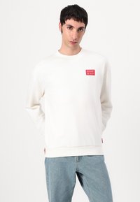 Kremet genser med rund hals, lange ermer og en rød logo på venstre bryst. Kombinert med lyseblå jeans.