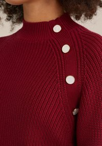 Burgundi spalvos megztinis su aukštu apykakle, su raukšlėta tekstūra ir dviem dekoratyviniais mygtukais ant peties, pridedančiais detalių.