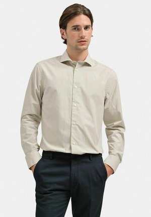 Lys beige skjorte med fine lodrette striber, lange ærmer og spids krave. Båret med mørke bukser, hænderne i lommerne.