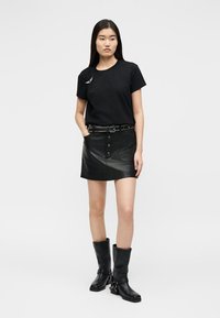 Γυναίκα που φοράει μαύρο T-shirt, μαύρη μίνι δερμάτινη φούστα με κουμπιά και ζώνη με τρουκς, καθώς και μαύρες δερμάτινες μπότες μέχρι τη μέση της γάμπας, στέκεται μπροστά σε λευκό φόντο.