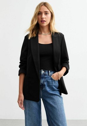 Svart kavaj med slag, två framfickor och uppvikta ärmar, kombinerad med svart linne och ljusblå vida jeans.