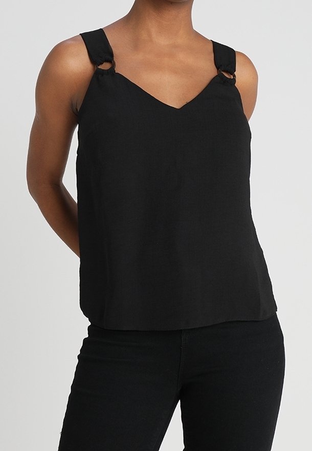 Femme portant un haut noir sans manches avec de larges bretelles ornées de détails en anneau, associée à un pantalon noir, sur un fond uni.
