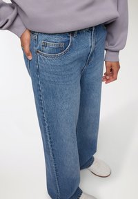 Jeans in denim blu chiaro a gamba larga presentano un classico design a cinque tasche e cuciture a contrasto, abbinati a un maglione grigio e a sneaker bianche.