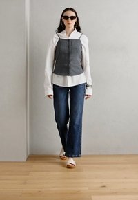 Gray fitted crop top over a white shirt, paired with flared blue jeans and white sandals. Model wearing sunglasses, simple hoop earrings.