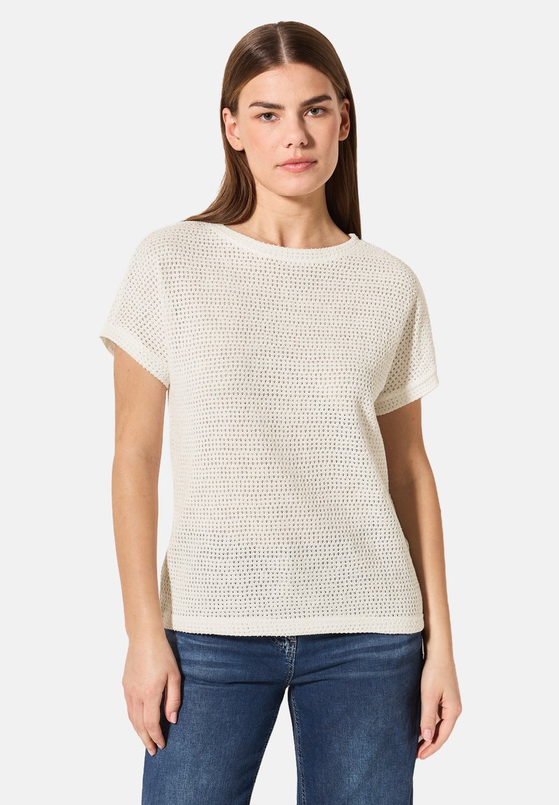 Lätt, krämfärgad stickad t-shirt med korta ärmar, rund halsringning och ett texturerat, öppet vävmönster. Matchas med blå jeans.