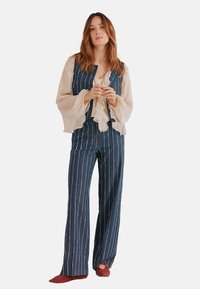 Tuta in denim con strisce verticali bianche, abbinata a una leggera blouse a balze. Presenta gambe larghe e un corpetto aderente. Scarpe rosse slip-on.
