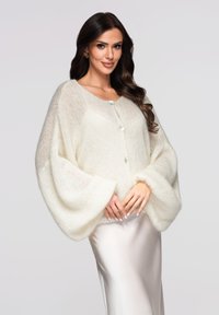 Cardigan surdimensionné de couleur crème fabriqué en tissu tricoté doux. Il présente de grandes manches, une fermeture à boutons et une surface légèrement texturée.