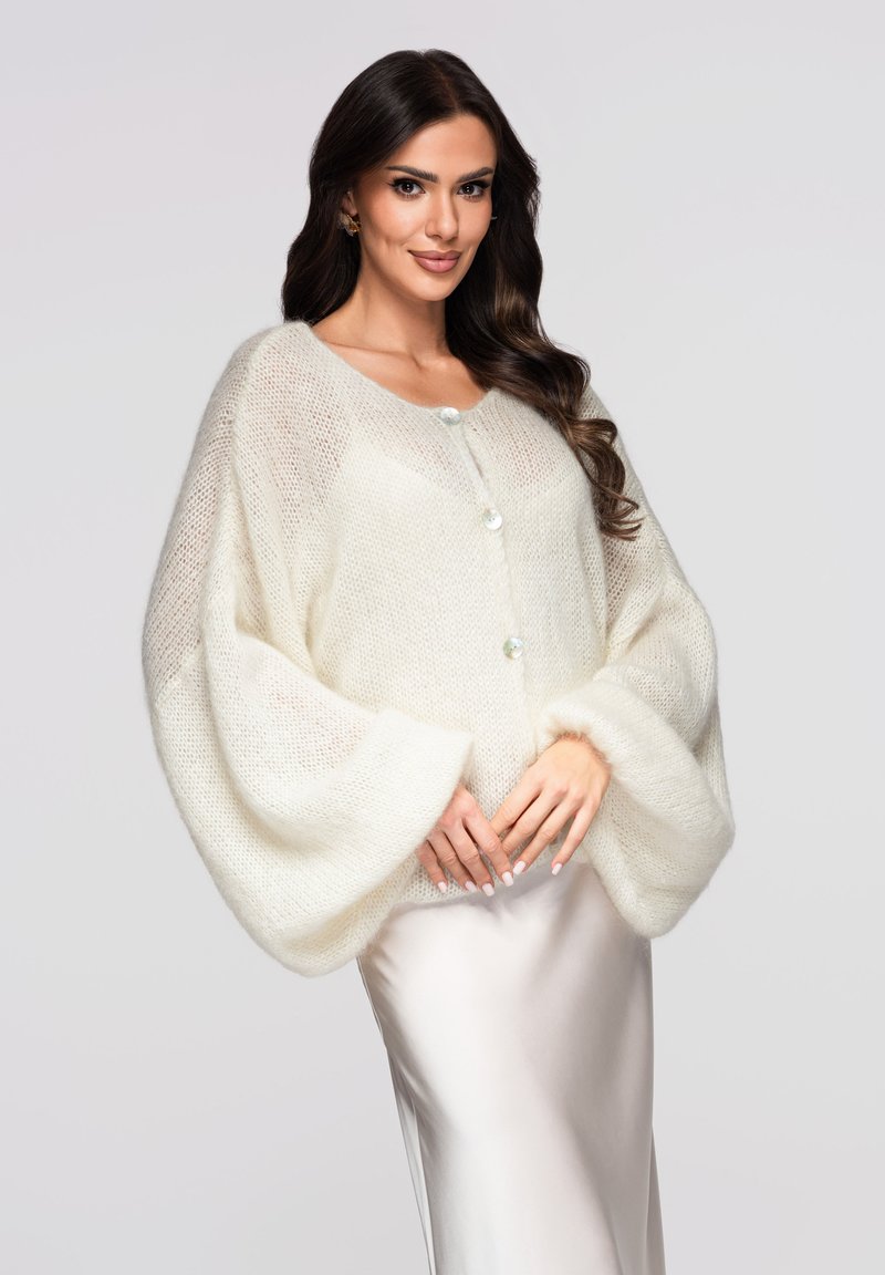 Cardigan surdimensionné de couleur crème fabriqué en tissu tricoté doux. Il présente de grandes manches, une fermeture à boutons et une surface légèrement texturée.