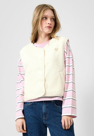 Jeune femme portant une veste en polaire crème boutonnée sur une chemise rayée rose et blanche à manches longues avec un jean bleu, regardant vers la droite.