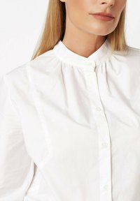 Woman wearing a white buttoned blouse with a high collar, showing her neck and lower face, and long straight blonde hair.