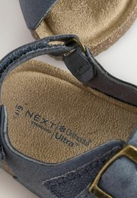 Hallid hallist seemisnahast sandaalid, korgist tallaga, reguleeritava rihmaga, kus on messingist luku ja tekstureeritud sisetald, millel on kiri "NEXT Standard/Ortholite Ultra."